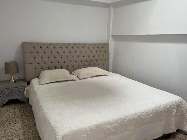 Una cama bien hecha con cabecera capitonada, dos almohadas y una mesita de noche con lámpara.