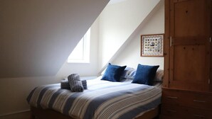 Ein Schlafzimmer mit schräg stehender Decke, einem Bett mit gestreifter Bettwäsche, blauen Kissen und einem Holzschrank.