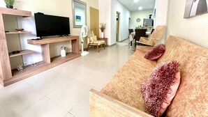 Un soggiorno con TV su un mobile in legno, un divano con cuscini decorativi, un'area pranzo sullo sfondo e un corridoio che conduce a un'altra stanza.