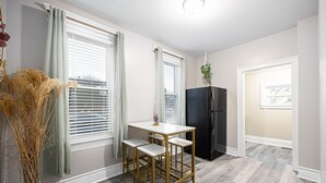 Eine Küche mit einem weißen Tisch und zwei Barhocker, einem schwarzen Kühlschrank und einem Fenster, von dessen Gardinenstange eine Pflanze hängt.