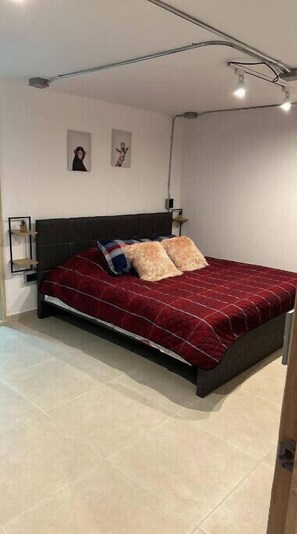 Quarto com cabeceira escura, colcha acolchoada vermelha, duas almofadas e uma pequena estante com uma moldura.