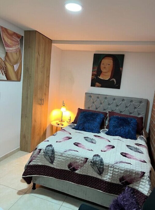 A bedroom with a tufted headboard, a bed with a patterned duvet, two blue pillows, a nightstand with a lamp, and a painting on the wall.