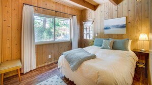 Un dormitorio con paneles de madera, una cama grande, una ventana con cortinas, un banco y una mesita de noche con lámpara.