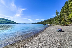 Ein Kiesstrand mit klarem Wasser, eine Reihe von Ferienhäusern und ein bewaldeter Hügel.