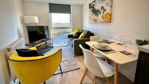 A modern living room with a TV, yellow chair, dining table with plates and glasses, and a sofa with yellow cushions. - Coates House one bed - Nailsea (Bristol)