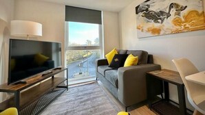 A modern living room with a flat-screen TV, a grey sofa with yellow cushions, a white chair, and a large abstract painting on the wall. - Coates House one bed - Nailsea (Bristol)