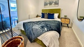 Ein modernes Schlafzimmer mit einem grünen, gepolsterten Kopfteil, einem großen Bett mit weißer und blauer Bettwäsche, einem kleinen Nachttisch mit einer Pflanze und einem großen Spiegel an der Wand.