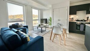 Uma sala moderna com um sofá azul, uma área de jantar com uma mesa branca e uma cozinha com armários escuros.