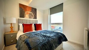 Um quarto com uma cama, mesa de cabeceira, luminária, janela e um quadro na parede.