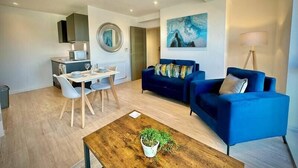 Uma sala de estar moderna com um sofá azul, uma mesa de centro de madeira com uma planta, e um espaço de jantar com uma mesa e cadeiras brancas.