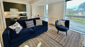 Een moderne woonkamer met een donkerblauwe bank, een bijpassende fauteuil en een salontafel. Er is een keuken met donkere kastjes en een magnetron. Een groot raam biedt uitzicht op een landelijk landschap.
