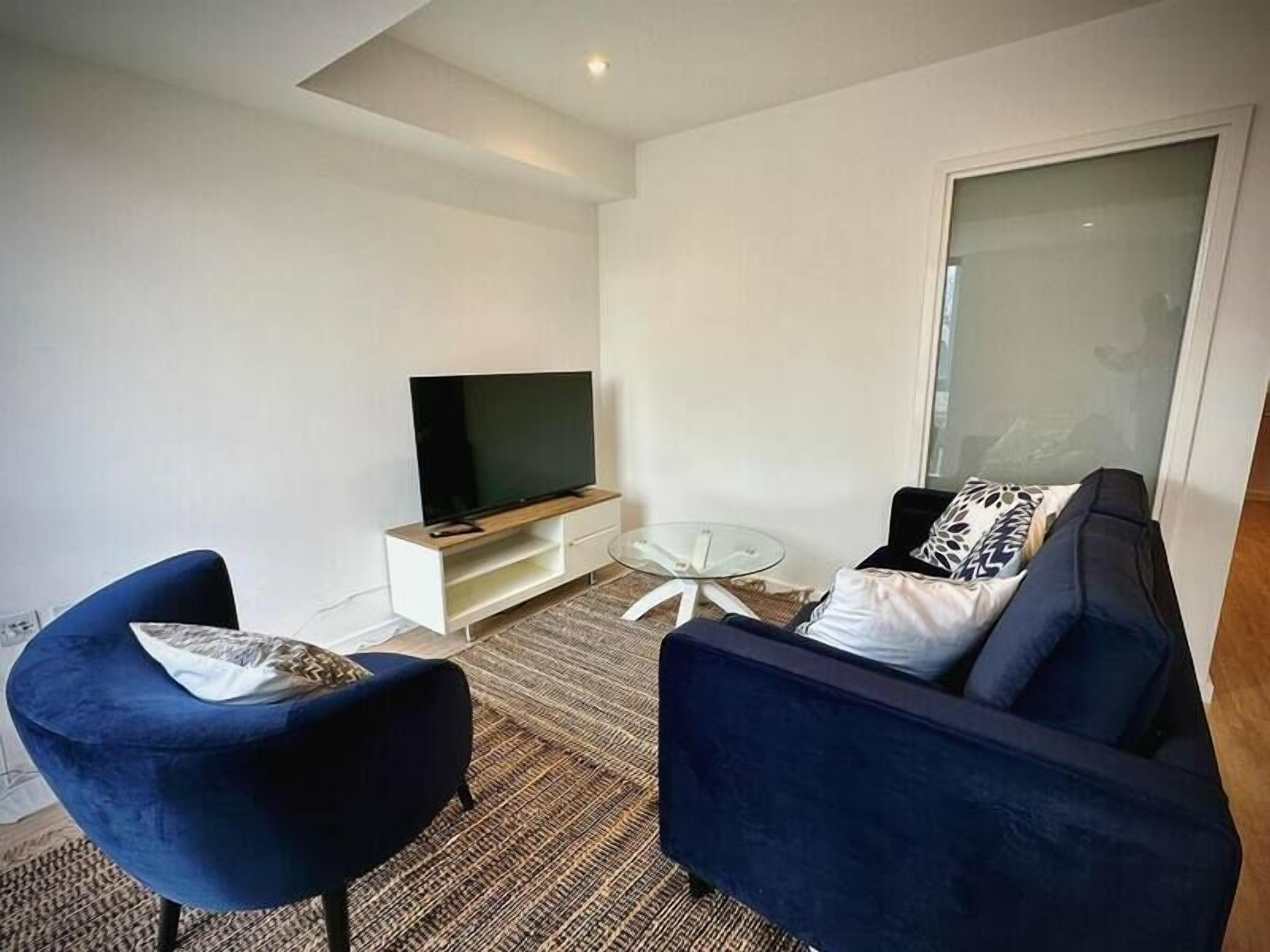Uma sala de estar moderna com uma TV de tela plana, duas poltronas azuis, uma mesa de centro de vidro e um tapete estampado.