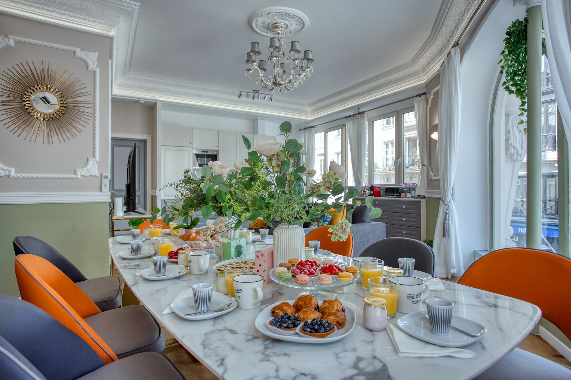 A breakfast setting with a marble table, orange chairs, and a variety of breakfast items including pastries, fruits, and beverages.