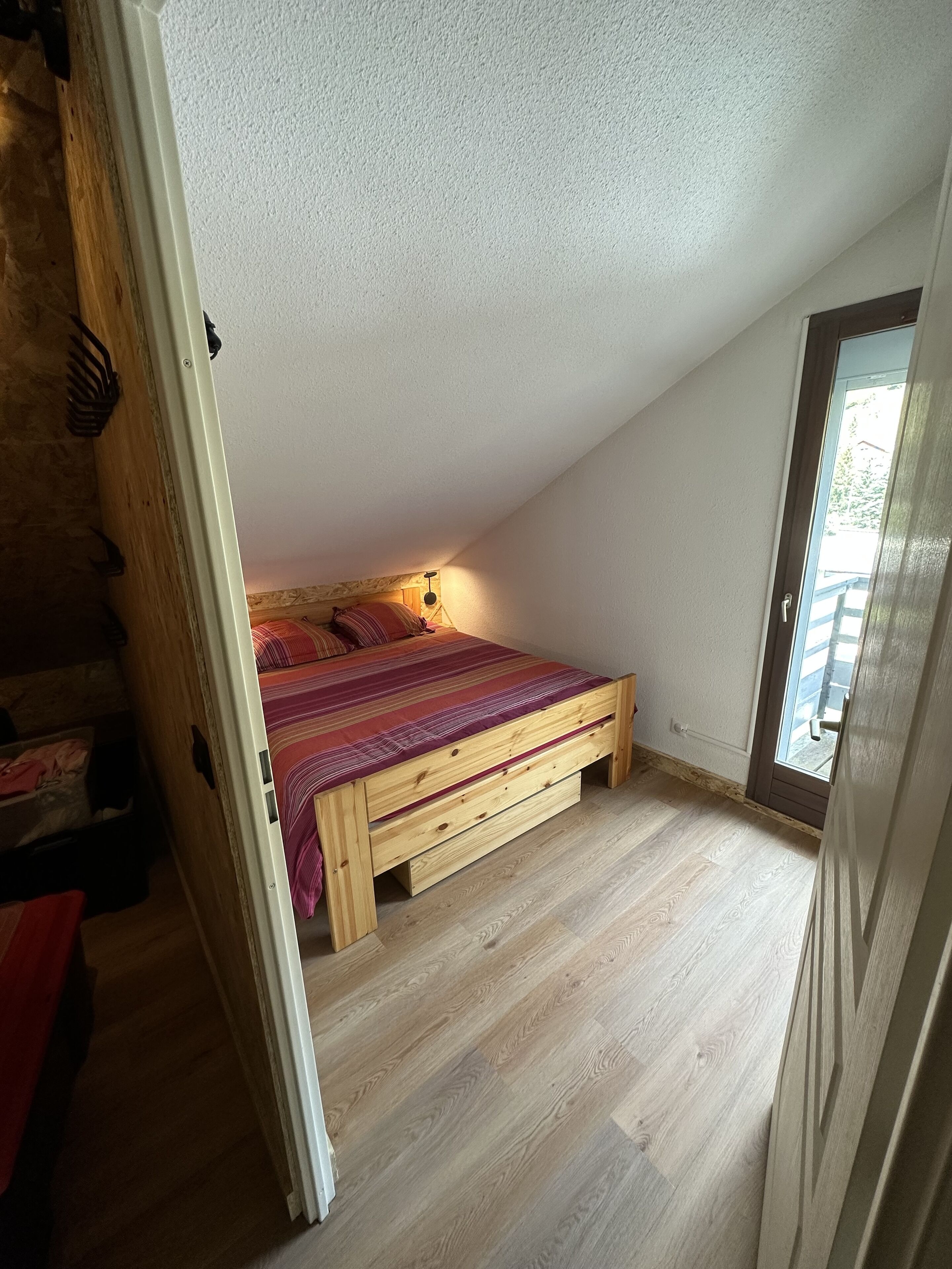 Une petite pièce avec un plafond en pente, un lit en bois, une petite fenêtre et une porte donnant sur une autre pièce.