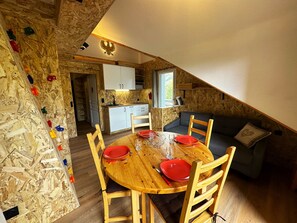 Un'atmosfera accogliente e rustica, con un tavolo da pranzo in legno apparecchiato per quattro persone, circondato da sedie in legno. Sullo sfondo, un divano grigio e una piccola finestra che si affaccia sul verde.