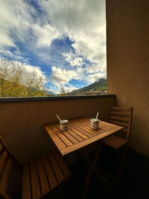 Un tavolo di legno con due tazze e una sedia, di fronte a un muro strutturato che offre vista su una montagna e alberi.