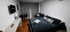 A modern hotel room with a bed, a desk, a chair, a TV, and a window with curtains. - Studio Centro Poços - Tudo a Pe (Poços de Caldas)