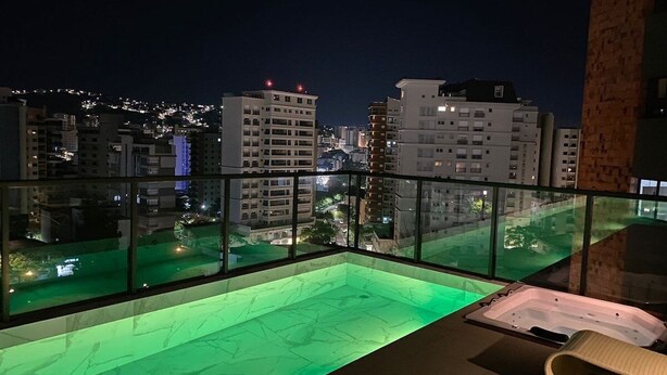 Una piscina en la azotea con barandilla de cristal, un jacuzzi y vistas de la ciudad iluminada de noche.