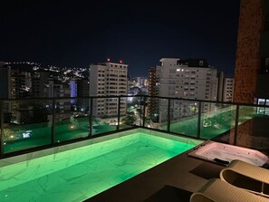 Una piscina en la azotea con barandilla de cristal, un jacuzzi y vistas de la ciudad iluminada de noche.