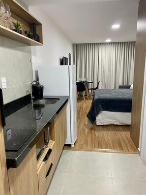 A compact hotel room with a kitchenette, a bed, a small table, and a curtain-covered window.