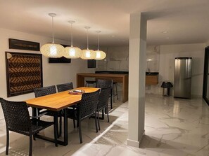 A modern dining area with a wooden table and black chairs, a kitchen with a refrigerator, and a patterned wall hanging. - Studio Centro Poços - Tudo a Pe (Poços de Caldas)