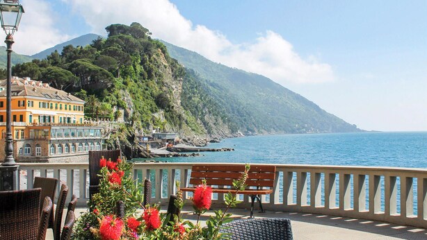 A coastal view with a bench overlooking the sea, a mountain in the background, and a building with a balcony.