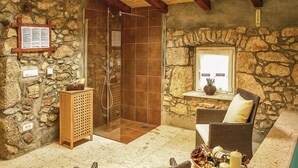 Un baño con paredes de piedra, una ducha con cerramiento de vidrio, un mueble de madera y una silla de mimbre con velas.