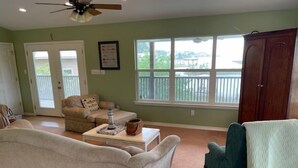 Un salon avec un canapé beige, un fauteuil vert et une table basse. Il y a une fenêtre donnant sur un balcon et une porte menant à l’extérieur.