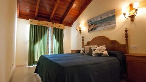 Quarto com teto de madeira, cama com colcha escura, duas luminárias na parede e um quadro de uma praia.