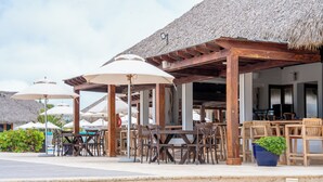 Un área de estar al aire libre con mesas y sillas de madera, sombrillas y una estructura de techo de paja.