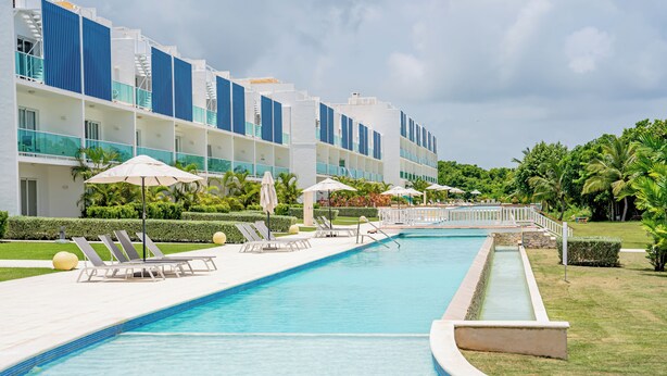 Una piscina con sillones de playa y sombrillas, rodeada por un edificio con varios balcones y un césped bien cuidado.