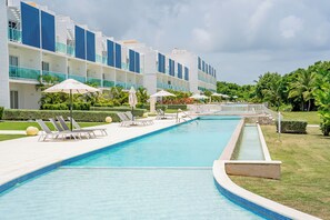A swimming pool with lounge chairs and umbrellas, surrounded by a building with multiple balconies and a well-maintained lawn.