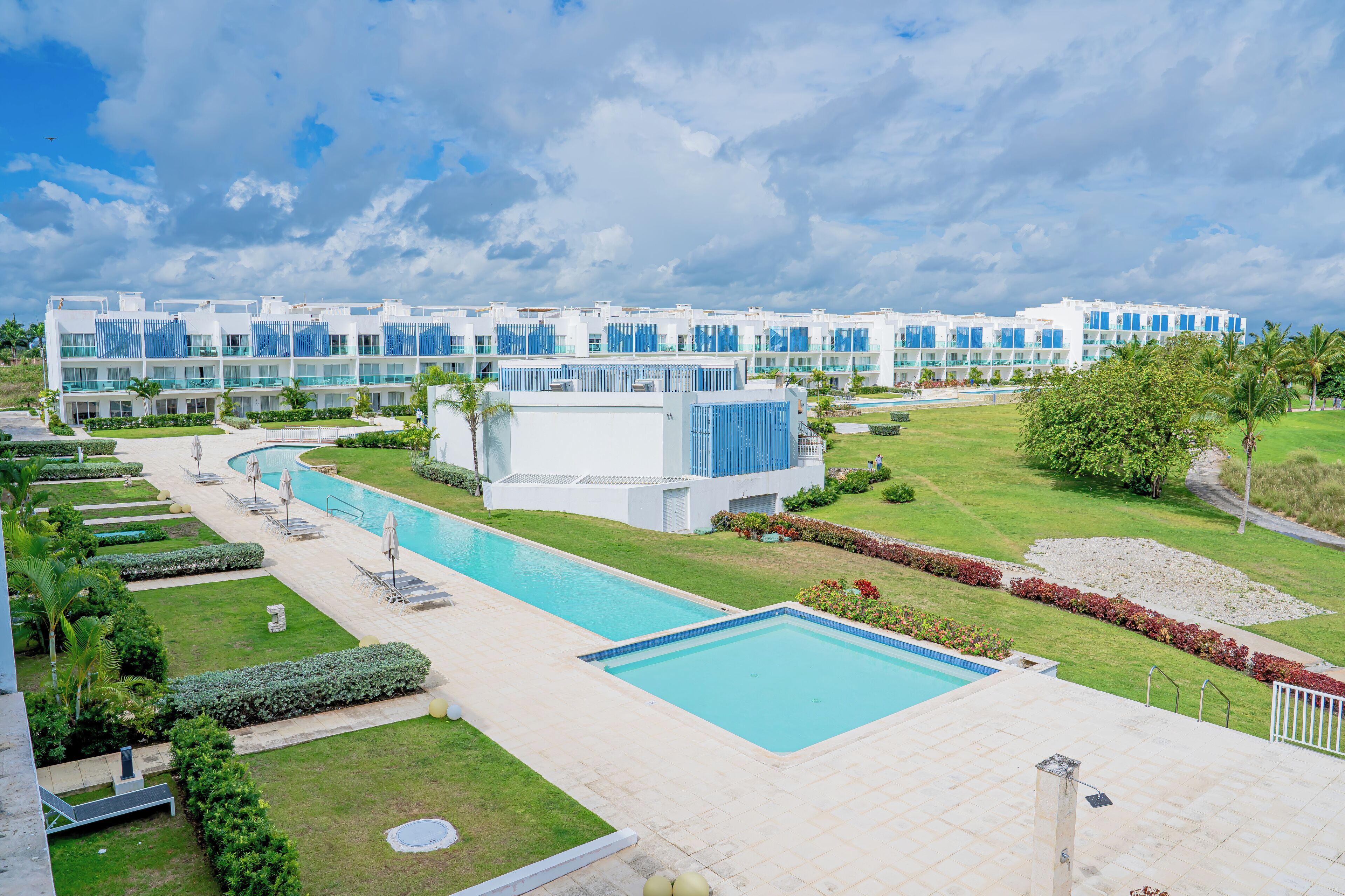 A modern hotel with a large swimming pool, well-maintained lawns, and a clear sky.