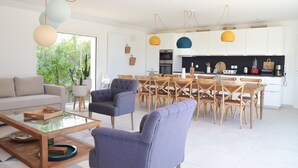 Un salon moderne avec un canapé, des fauteuils et une table basse. On aperçoit également un coin repas avec une table et des chaises en bois.