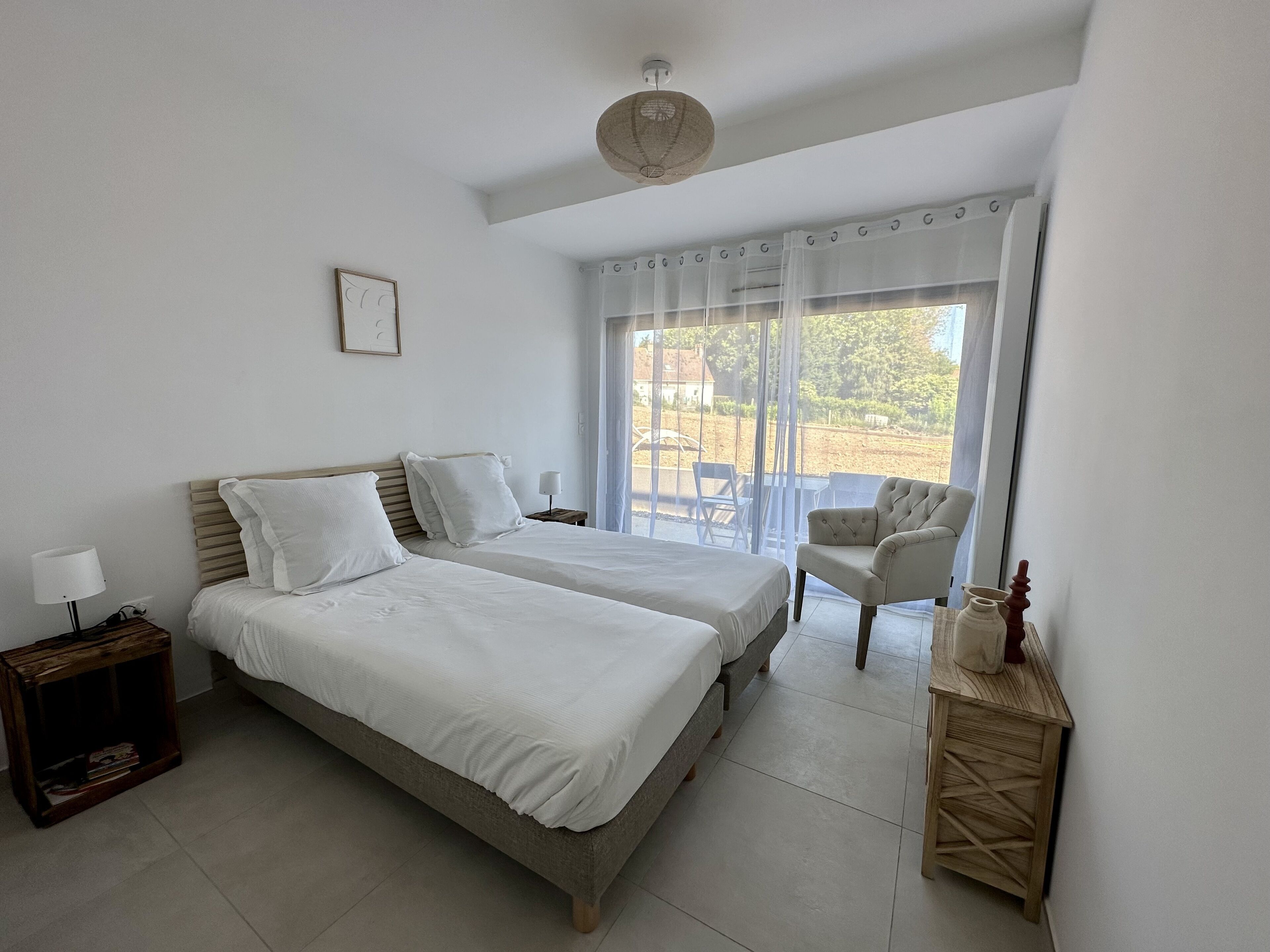 A bedroom with two beds, a chair, a table, and a view of the outdoors.