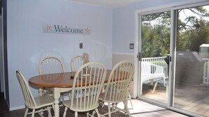 Un coin repas avec une table ronde et quatre chaises, un panneau « Bienvenue » accroché au mur et une porte-fenêtre coulissante donnant sur une terrasse extérieure.