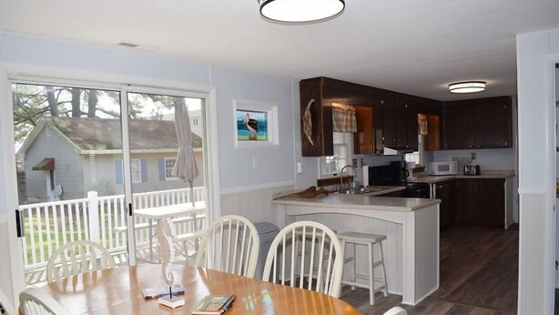 Un coin repas avec une table en bois et des chaises blanches, une cuisine avec des meubles sombres et un îlot central blanc, et une vue sur une maison à travers une porte vitrée.
