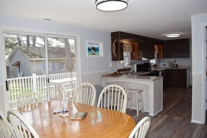 Un coin repas avec une table en bois et des chaises blanches, une cuisine avec des meubles sombres et un îlot central blanc, et une vue sur une maison à travers une porte vitrée.