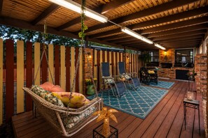 Een houten terras met een hangstoel, twee loungestoelen, een bakstenen openhaard en een stenen muur.
