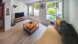 A living room with a TV, a wooden coffee table, a sofa, and large windows with a view of trees and a balcony. - 3 bedroom beach front apartment in Agde (Agde)