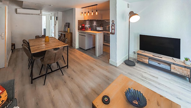 A modern living room with a wooden dining table, a TV on a wooden stand, and a floor lamp. - 3 bedroom beach front apartment in Agde (Agde)