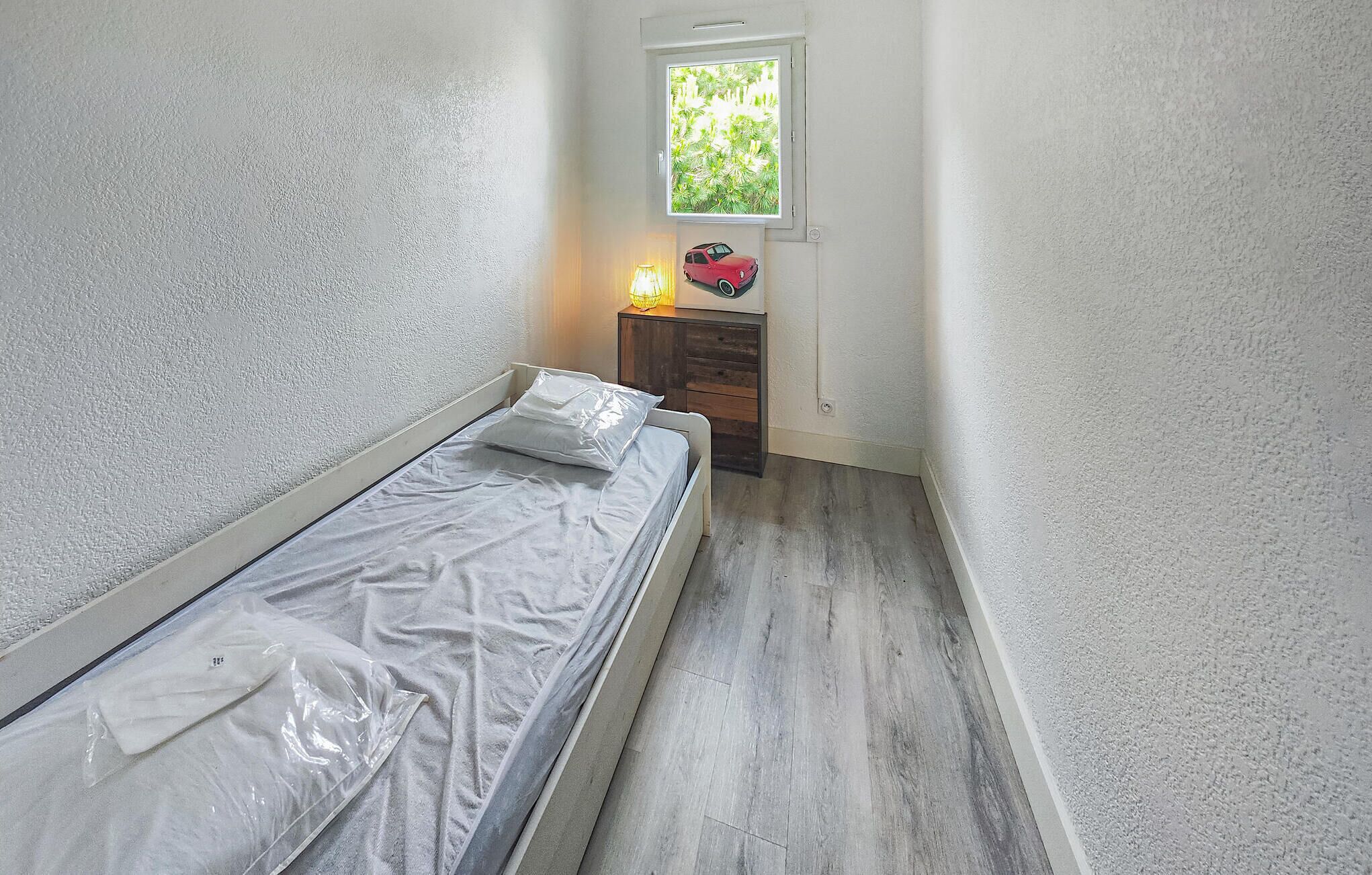 A small, single-bed room with a wooden floor, a window with a view of greenery, a bedside table with a lamp, and a wall-mounted picture.