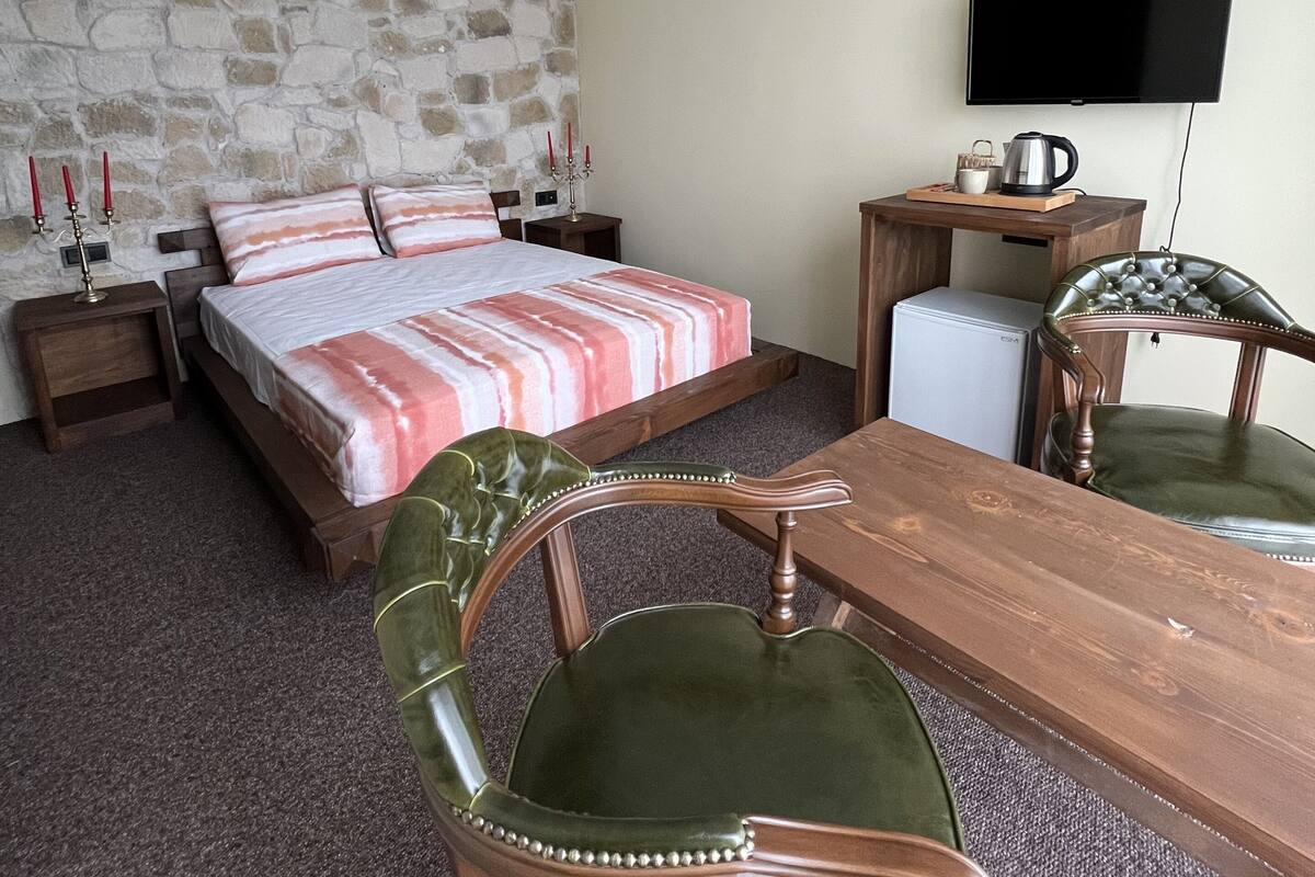 A hotel room with a bed, a wooden table, a green leather chair, a television, and a stone wall.