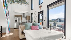 Un dormitorio moderno con una cama, un mural de paisaje urbano, una cocina al fondo y un amplio ventanal con vistas al exterior.