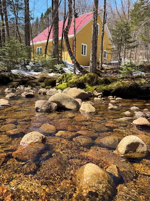 Un ruisseau aux eaux claires, bordé de rochers, entouré d’arbres et avec, à proximité, un bâtiment jaune doté d’un toit rouge.