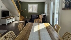 A spacious living room with a large wooden dining table, a sofa, and a TV. A staircase leads to an upper level. - 5 minutes from the Bras du Nord Valley / Chalet with riverfront spa (Saint-Raymond)