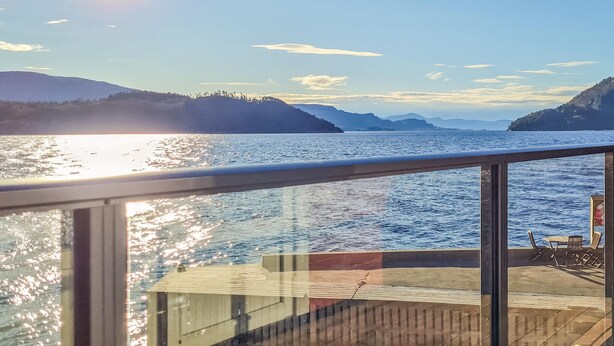 Vue depuis le pont d’un bateau, avec une rambarde en verre, un pont en bois et une table entourée de chaises.