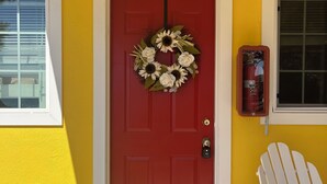 A red door with a floral wreath, a white chair with a red and white checkered cushion, and a welcome mat with a pickup truck design. - ALL INCLUSIVE AMENITIES:Kayak,SUP,Pedal Boat,PingPong,Pickleball,Movie Nt &MORE! (Nice)
