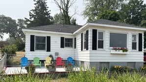 Ein weißes Haus mit schwarzen Fensterläden, eine Veranda mit bunten Adirondack-Stühlen und ein Garten im Vordergrund.