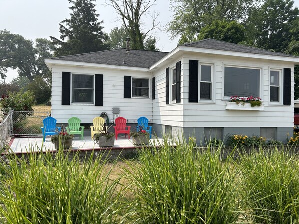 Una casa blanca con contraventanas negras, un porche cubierto con sillas Adirondack de colores y un jardín en primer plano.